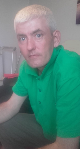 Image shows a man. He has fair skin and blue eyes. His hair is fair and cur short. He is wearing a green t-shirt with three buttons and a collar.