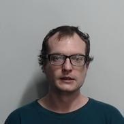 A head and shoulders image of a white man in front of a white background. The man has short brown hair and is wearing black rimmed glasses and a dark green t-shirt.