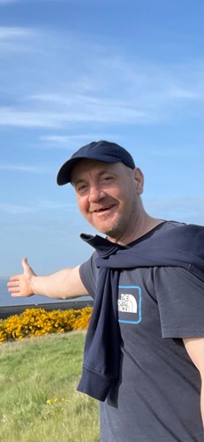 Image shows a man. He is standing outside, with a clear blue sky behind him. He is wearing a dark blue cap, a dark blue t-shirt and a dark blue jumper, which is draped over his shoulders. He is smiling broadly and gesturing behind him with one arm.