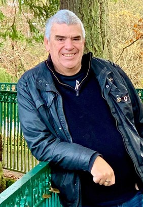 Image of man who died, Andrew MacPherson, smiling man with short grey hair, dark eyebrows, wearing dark-coloured wax or leather jacket, and dark-coloured pullover. Leaning on green fence in front of conifer tree.