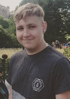 Young man with short hair standing in a garden. He is wearing a grey coloured top and is smiling at the camera. There are trees and people behind him.