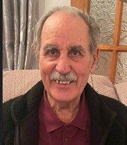 85-year-old white man with grey hair and moustache wearing a burgundy collar shirt and black jacket