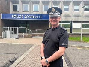 Male police officer wearing black uniform and hat standing in front of Police Scotland sign