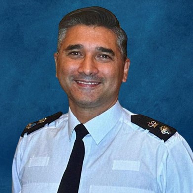 Ch Supt Faroque Hussain smiling in front of a blue background. He is wearing a white police shirt and black tie and epaulettes with a pip and a crown.