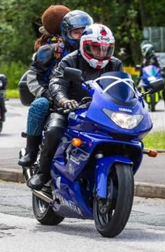 Two people on a blue motorcycle travelling down a road with other motorcycles in the background