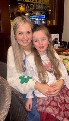 Image of two females. One is a child sitting on the other woman's lap. The woman has long blonde hair and is wearing a white jumper. The girl also has long hair and is wearing a white top and red skirt with a love heart design.