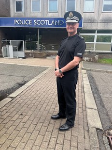 Male police officer wearing black uniform and police hat standing in front of a Police Scotland sign