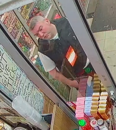 White man leans over counter in shop with grey hair and black body warmer