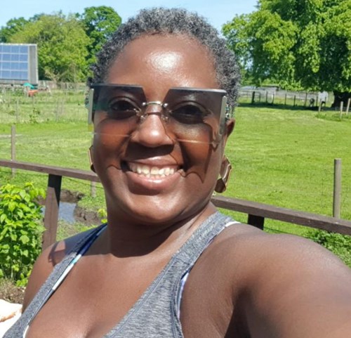 Image of woman who died Chitsidzo (Veronica) Chinyanga - smiling black woman with short greying hair wearing grey vest top and square sunglasses in front of field.