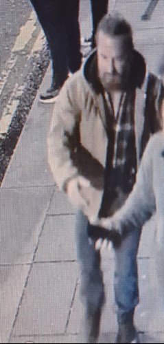 white male, around 35-40-years-old, stocky build with dark hair worn in a bun and a beard. Wearing a tan coloured jacket, checked shirt and blue jeans.
