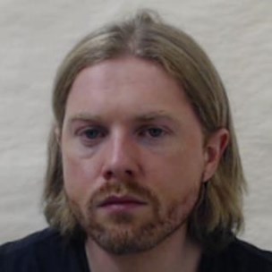 White man with blond hair in a middle parting. He is looking straight at the camera and has beard growth. He has his hair tucked behind his ears and he is wearing a black top