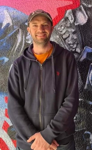A smiling man, wearing a baseball cap. He has slight facial hair and is wearing an orange t-shirt below a dark blue, zip-up hoodie.