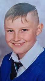 Head and shoulders image of white nine-year-old boy. Short brown hair with shaved sides, brown eyes. Wearing a white shirt with a black tie with blue, white and green stripes on it and a blue jumper on top