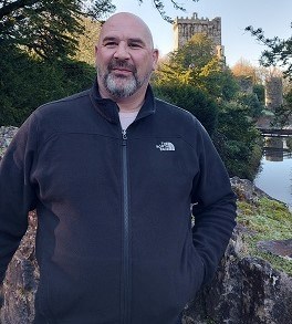 50-year-old man standing on a bridge over water with trees and a building in the background