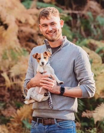 Image of man who died, Cameron Pearson. Smiling, wearing a grey jumper, jeans and brown belt, holding dog  in sunlight with ferns in the background