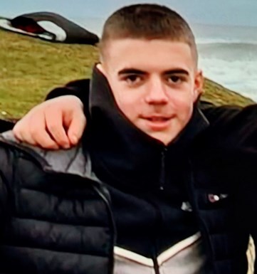 Image of missing teenager Kayden Thompson - male teenager with short brown hair, dark eyebrows, brown eyes, wearing black grey and white zip-through top and black puffer jacket standing on cliff with sea behind him.