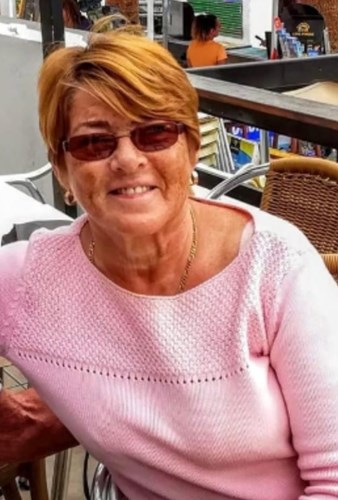 Image shows a woman. She has short auburn hair and is wearing sunglasses and a pink jumper. She is smiling.