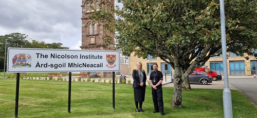 Two people stood next to a sign that says the Nicolson Institute. One is male and is a police officer in uniform. The other is female with long blonde hair and wearing a dark coloured suit. They are standing next to a tree and the school building can be seen behind them.