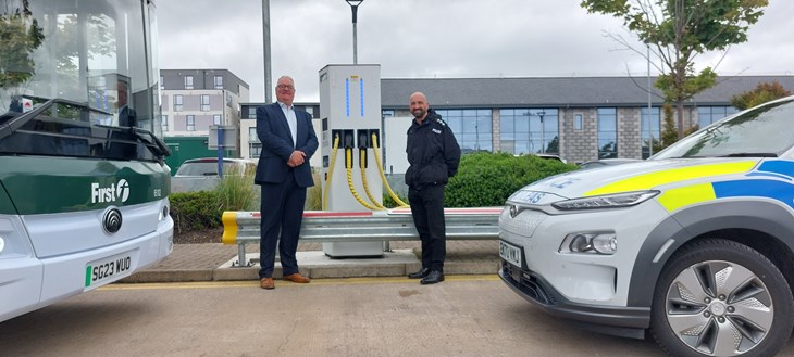 First Bus' David Adams & CI Darren Bruce stand at an electric vehicle charging point between an electric bus & marked police car.