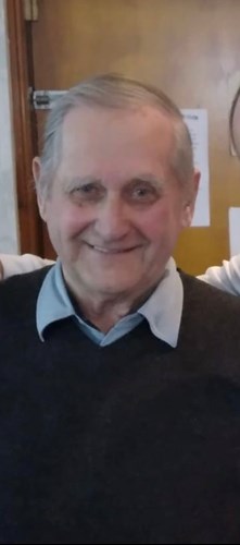 Image shows an older man. He has grey hair and is smiling broadly at the camera. He is wearing a dark v-neck jumper and a blue shirt with a collar.