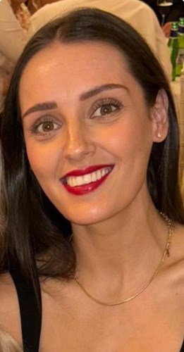 Image shows a young woman. She has dark brown hair and brown eyes. She is wearing red lipstick and is smiling broadly.