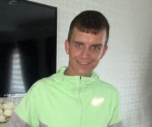 Young man with short dark hair, standing in front of a TV and a white wall. He is wearing a bright green top with a white Nike logo. He is staring straight ahead and smiling.