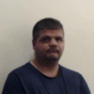 White man with very short dark hair. He is staring straight into the camera and is wearing a navy blue t-shirt. He is standing in front of a white wall.