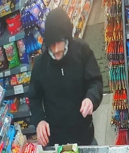 Images shows a man. He is pictured in a shop. He is wearing dark clothes with his hood pulled up.