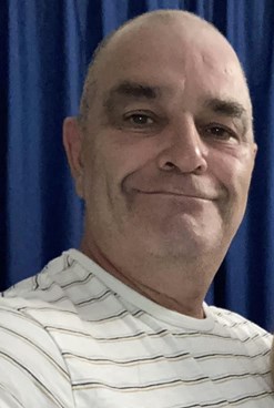 Image of Keith Rollinson who died following assault in Elgin. Smiling bald man with white striped tshirt and blue curtain behind