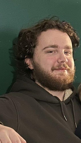 Image shows a man. He is in front of a green background. He has fair skin, curly hair and a beard.